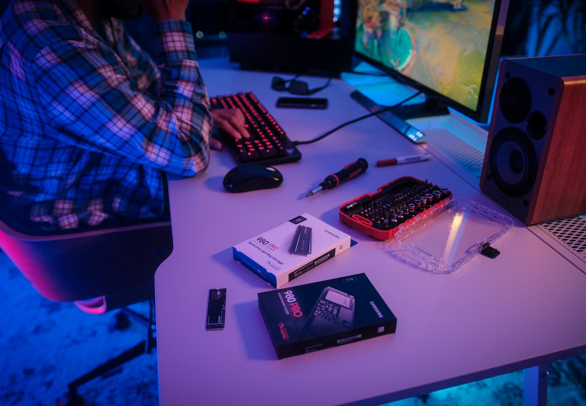 Person sitting at a gaming PC desk at night with a Samsung 980 PRO SSD, screwdriver kit, and tools laid out — preparing for a PC hardware upgrade or diagnostic