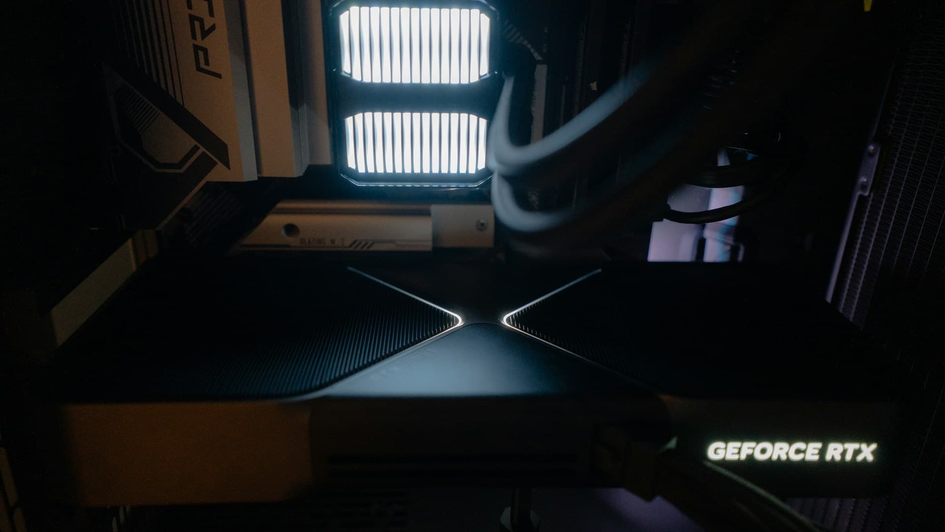 Interior of a PC case showing an NVIDIA GeForce RTX graphics card with illuminated X-shaped lighting on the shroud, lit case fans in the background, and white cable management visible
