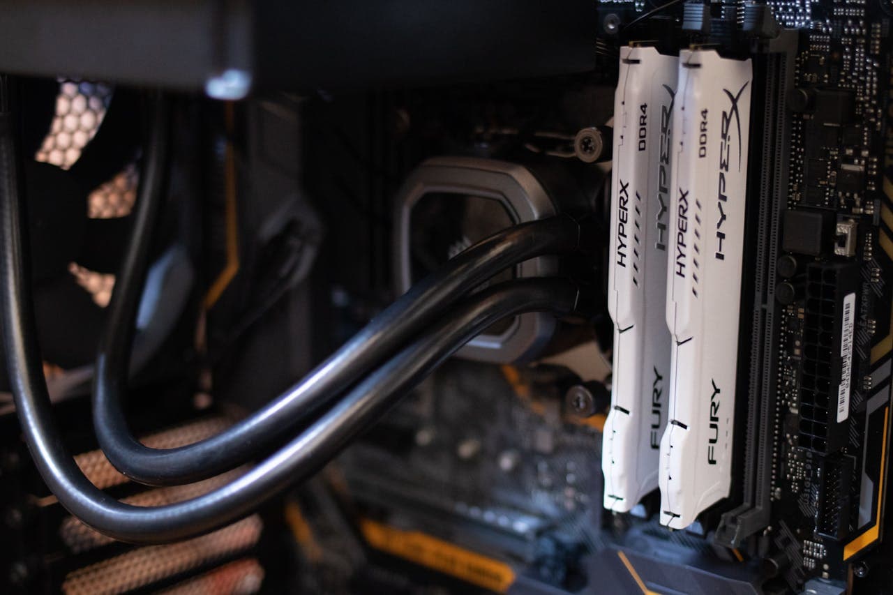 Interior of a desktop PC showing two white HyperX Fury DDR4 RAM sticks installed on a motherboard, with liquid cooling tubes and a case fan visible in the background.