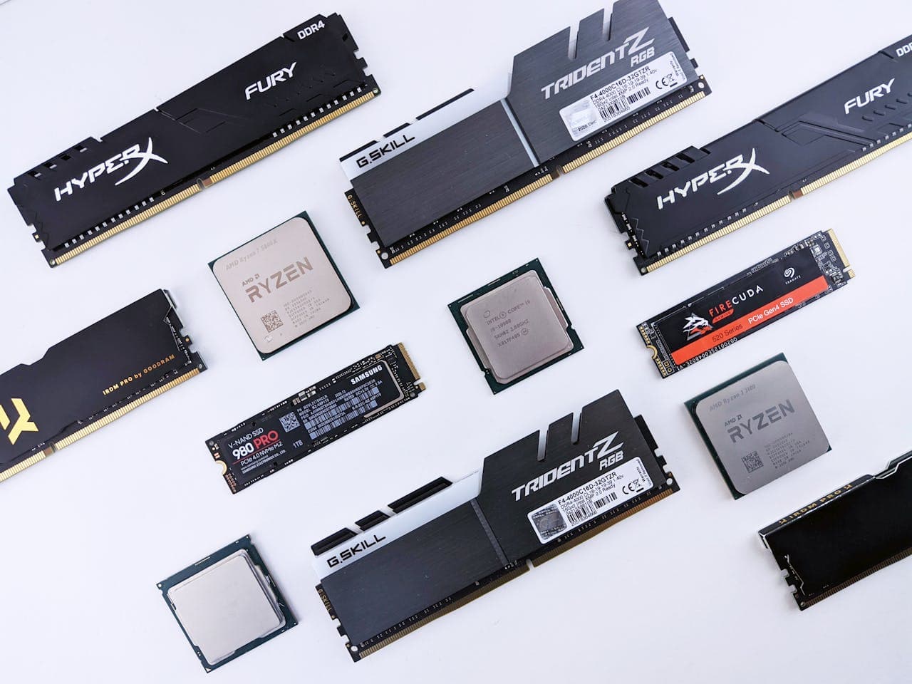 Flat-lay of PC upgrade components on a white surface including HyperX and G.Skill DDR4 RAM sticks, Samsung 980 Pro and Seagate FireCuda NVMe SSDs, AMD Ryzen and Intel Core CPUs arranged in a knolling style